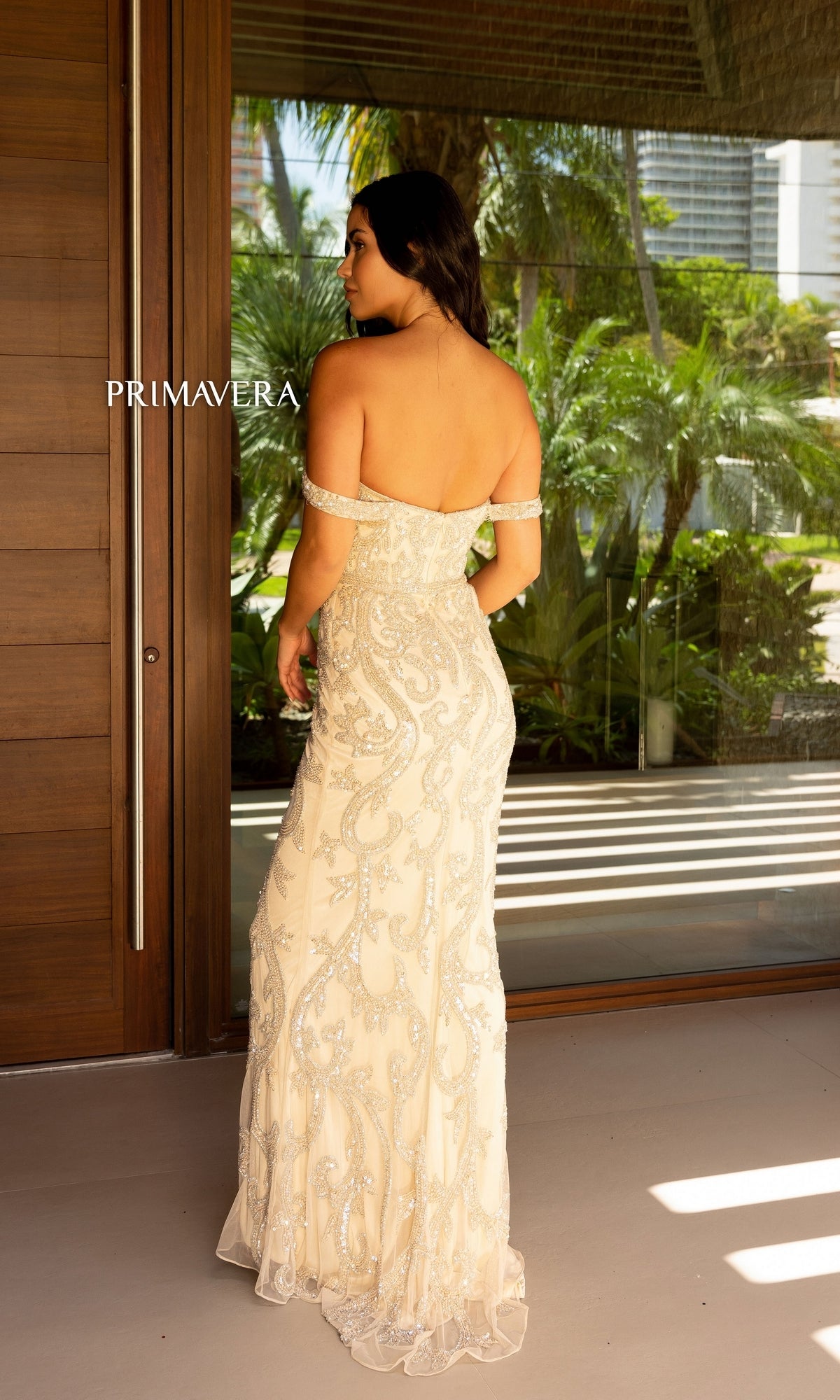 A woman stands facing away in front of a glass door, wearing the Primavera Beaded Off-the-Shoulder Prom Dress 12110—its floor-length cream fabric and silver embellishments softly glowing, with greenery visible outdoors.